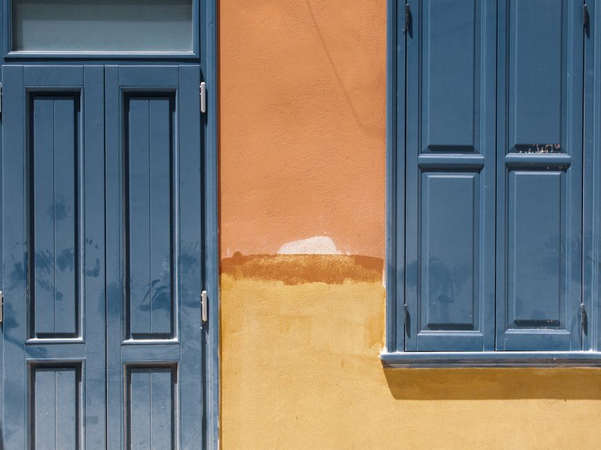 Vibrant orange wall with a blue door and window in daylight, showcasing contrasting colors.